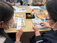 長い茶色のテーブルの上で、顔を寄せ合う二人の中年女性が、手元にある細長い木製のような飾りと小さなペンチを使い、熱心に手芸作業に取り組む近接写真