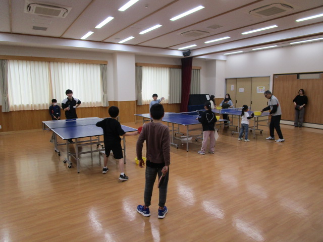 照明が明るいフローリングの広間には3台の卓球台が並べられ、子供たちと大人たちがそれぞれラケットを持って、和やかな雰囲気の中で卓球を楽しんでいるスポーツ活動の様子を捉えた写真