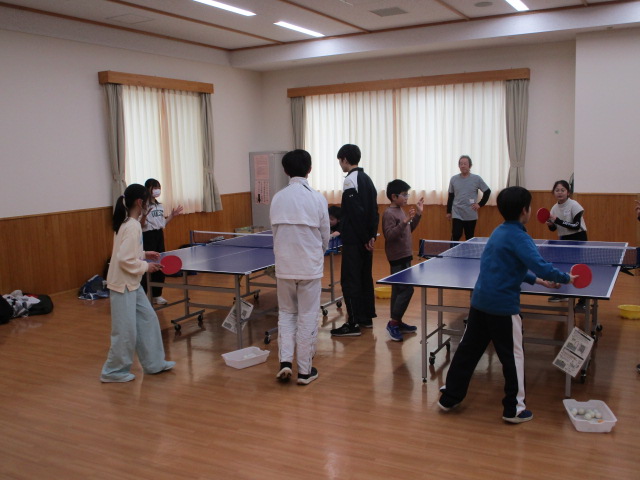 大きな窓のあるフローリングの広間には卓球台が2台設置されており、手前と奥で子供たちや大人がそれぞれ卓球のラリーや練習を楽しむ様子が写されている、スポーツ交流会の風景写真