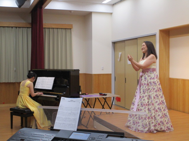 ピアノと電子キーボードが配置された舞台のような室内で、右側には花柄のロングドレスを着た女性歌手が笑顔で歌い、左奥では黄色のロングドレスを着た女性ピアニストが黒いアップライトピアノを演奏しているコンサートの様子を捉えた写真
