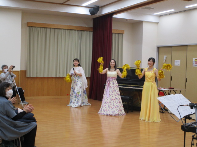 白い花柄のロングドレスを着た女性2人と黄色いロングドレスを着た女性1人が、中央の黒いピアノの周りで金色のポンポンを持って笑顔でパフォーマンスを披露しており、観客が拍手しているコンサートの様子を捉えた写真