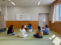 窓際の日当たりの良い和室で、畳の上に座った大人と幼児が、中央にいる青い服の女性講師による絵本の読み聞かせを聞き、後方のホワイトボードには平仮名やイラストが貼られている子育て支援イベントの様子を捉えた写真