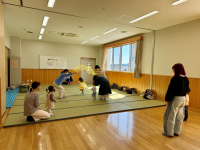 木張りの床と畳スペースを持つ広間で、保護者やスタッフと思われる複数の大人たちが、手や体を使った遊びで歩き始めたばかりの幼児たちと交流し、奥には明るい窓とホワイトボードが見える子育て支援イベントの様子を捉えた写真