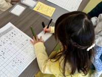 三つ編みの髪の毛をした黄色い服の幼い子供が、右手にペンを持って、小さなメモ帳に何かを書き込んでいる様子を上から捉えた写真