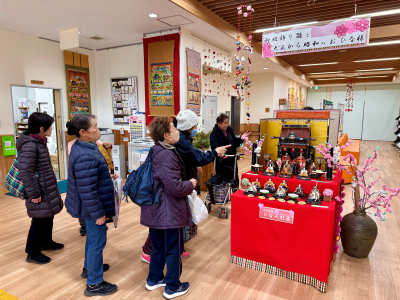 赤い布を敷いた台に飾られた雛人形を、厚手の冬服を着た女性たちが熱心に見学しており、右奥の天井から吊るされた飾りや、背後の壁に飾られたタペストリーのようなものも見える展示風景の写真