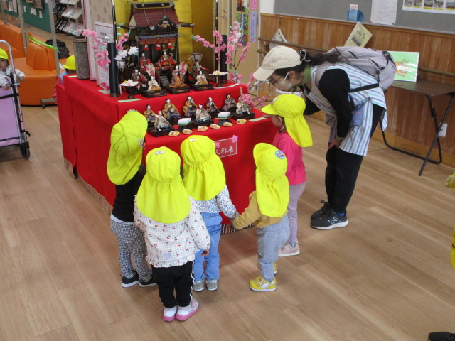 台の上に雛人形が飾られ、その周りに黄色い帽子をかぶった園児たちが集まり、かがみ込んだ保育士が優しく説明している様子を、保育室のような空間で捉えた写真