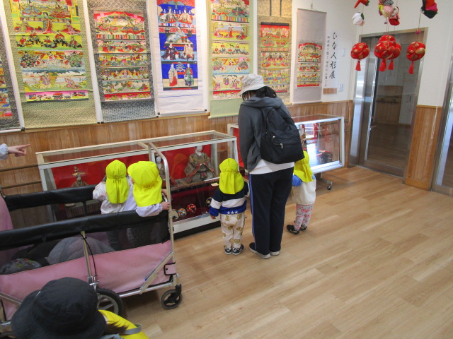 施設のフローリングの部屋で、保育士と思われる女性が引率する黄色い帽子を被った幼い園児たちが、ガラスケースの中に展示された豪華な段飾りの雛人形を鑑賞している様子の写真