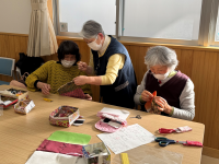 施設の明るい部屋のテーブルで、マスクを着用した3人の女性たちが、それぞれ布製の小物を手縫いで作っており、一人がもう一人に身を乗り出して手元を教えているような交流の様子を捉えた手芸活動の写真