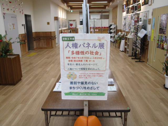 長テーブルの中央に白地のパネルが置かれ、令和6年度「人権パネル展」「多様性の社会」のタイトル、令和7年3月16日から29日までの期間、会場は西公民館1階ホールと記された啓発展示の写真