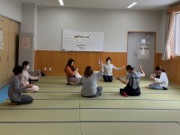 和室のような部屋で、4人の女性が子どもを抱っこして畳の上に円形に座って手を広げるポーズをして、手あそびをしている写真