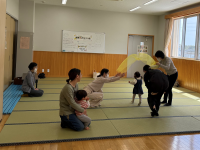 和室の広間で、数人の母親と幼児たちが、黄色い布を持ってくぐり抜けて遊ぶ姿や抱っこや見守りといった交流をしている写真