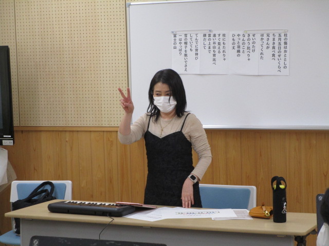 木目調の壁の部屋で、黒いキャミソールワンピースを着てマスクを着用した女性が、電子キーボードが置かれた机の前に立ち、ホワイトボードの童謡らしき歌詞を背に、ピースサインをしている様子の写真