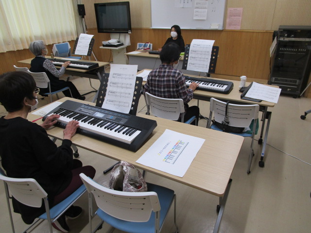 木目調の壁の部屋で、4人の生徒がそれぞれ譜面台付きの電子キーボードに向かって座っており、正面の講師らしき人物の指導で演奏している写真