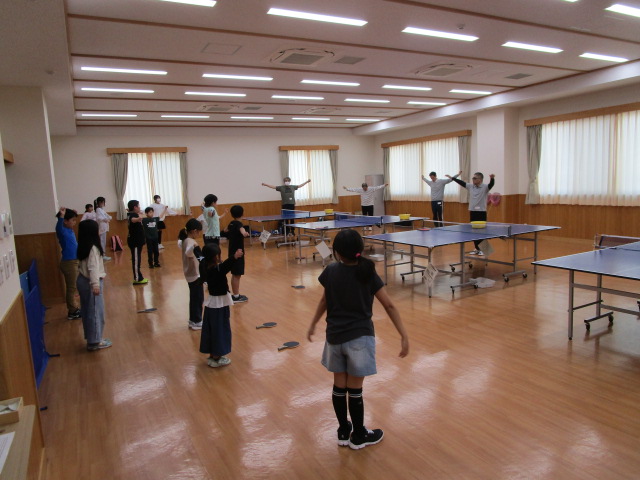 明るい板張りの広い室内で、約10人の子どもたちと数人の大人が卓球台の周りに立ち、一斉に両手を広げて準備体操をしている様子の写真