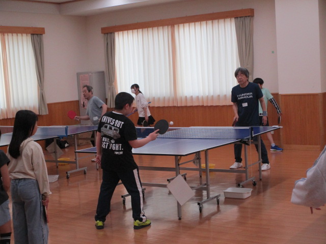 明るい木目調の床の体育館のような部屋で、手前の卓球台で黒いシャツの男の子が大人の男性と真剣にラリーの練習をしており、奥の卓球台でも別のペアが卓球を練習している様子の写真