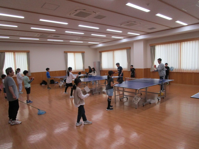 卓球台が複数設置された広い室内で、それぞれの卓球台に子どもたちが並んで、対面の指導者の大人から卓球の練習や指導を受けている様子の写真