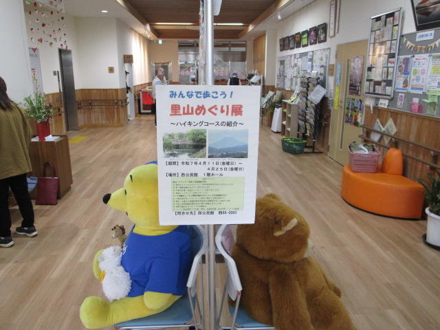 施設の明るいフローリングの廊下に、プーさんを模した大きな黄色いぬいぐるみと茶色いぬいぐるみが座っているスタンドにハイキングコースを紹介する「みんなで歩こう！里山めぐり展」のポスターが掲示された写真