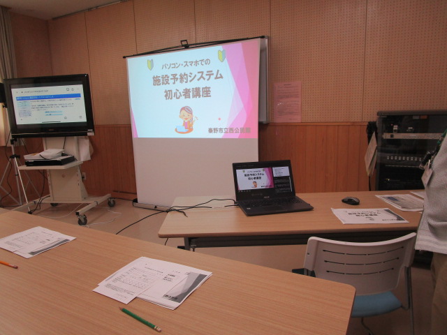 中央のスクリーンに映し出された講座タイトルと、左のモニター、手前の机上の資料やノートパソコンが置いてあるパソコン・スマホの施設予約システムの初心者講座の会場風景の写真