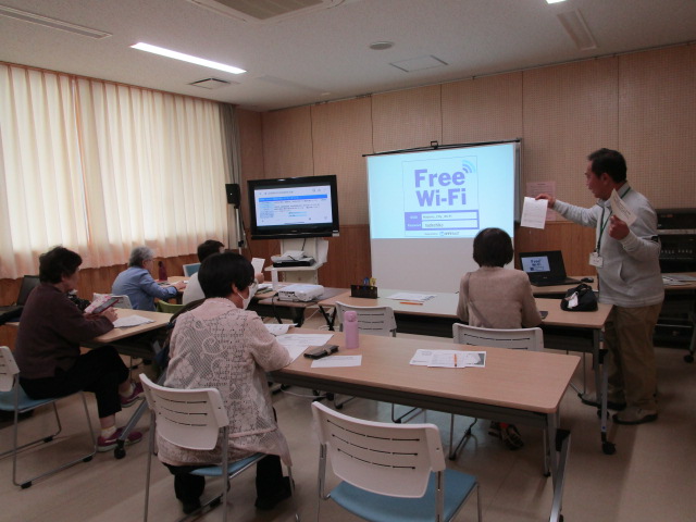 公民館のような部屋で、Free Wi-Fiの接続画面がスクリーンに投影されている前で、講師らしき男性が資料を手に説明し、数名の受講生が熱心に話を聞いている予約システム講座の様子の写真