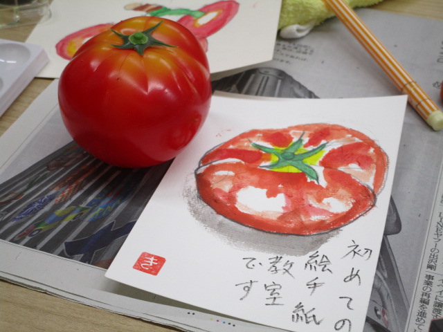 テーブルの上に置かれた本物そっくりのトマトの食品サンプルと、それをモチーフに描かれた、水彩画のようなタッチの「初めての絵手紙 教室です」と手書きされたトマトのイラスト入りの葉書の写真