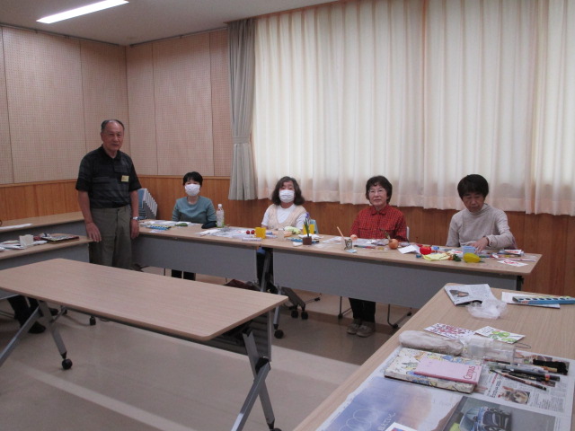 公民館のような部屋で、絵手紙で使用する道具が広げられた長机に座った4人の女性と立っている講師らしき男性がカメラに向かって穏やかな笑顔を見せている写真
