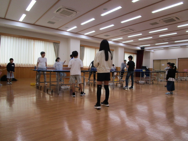 体育館のような広い部屋で、複数設置された卓球台に子どもたちが並び、向かい側に立つ大人と卓球の練習をしている写真