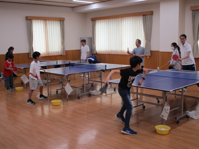 体育館のような広い部屋で、手前の卓球台でラケットを振る男の子を中心に、複数の卓球台で子どもたちと大人が卓球の練習をしている様子の写真
