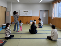 畳敷きの広い部屋で、数組の親子が幼い子どもを抱っこして座ったり立ったりして絵本の読み聞かせを聞いている写真