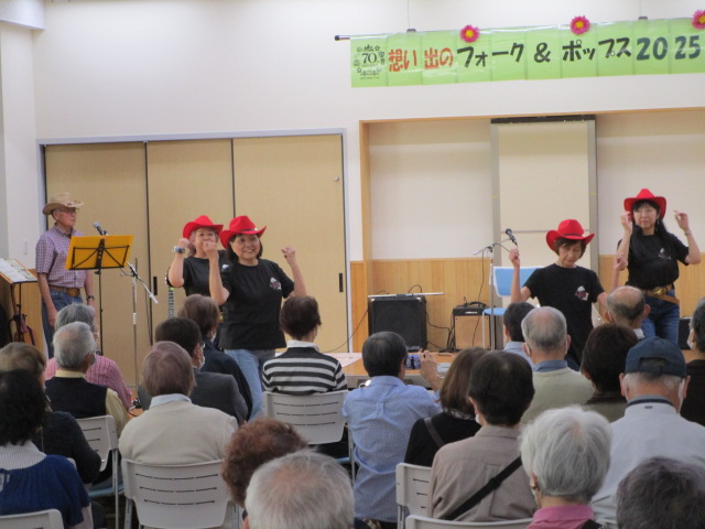 屋内ステージ上で、赤色のカウボーイハットと黒いTシャツ姿の4人の女性が、カントリーミュージックに合わせて踊りを披露しており、多くの観客が白い椅子に座って鑑賞している様子の写真