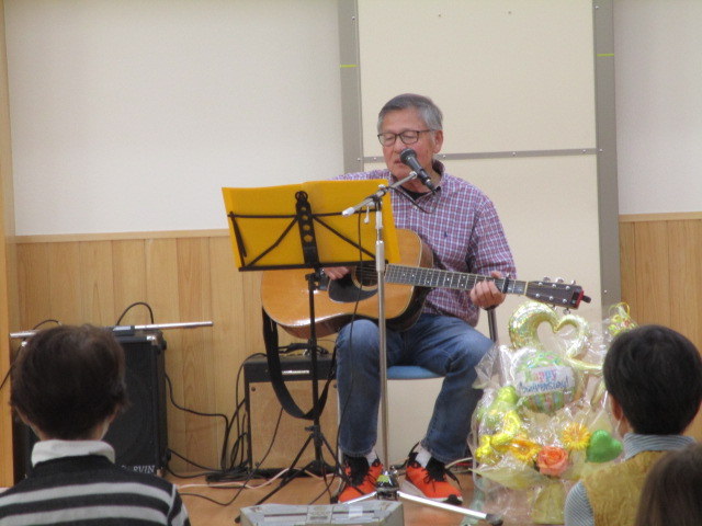 屋内ステージで、チェックシャツにジーンズ姿の年配男性が椅子に座ってアコースティックギターを弾きながら歌を披露している写真