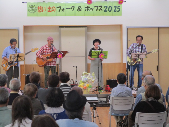 「70's 想い出のフォーク＆ポップス 2025」と書かれた横断幕の下で、3人の男性がギターを弾き、1人の女性がマイクスタンドの前で歌うバンドが、多くの観客の前で演奏しているライブの様子の写真