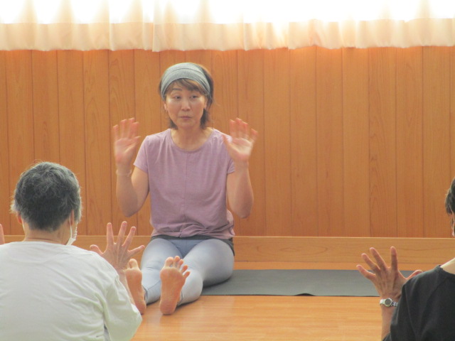 木目調の壁を背にした室内で、ヨガマットの上に座った女性講師が、両手を上げて身振り手振りを交えながら、座っている生徒たちにストレッチや体操の方法を説明している様子の写真