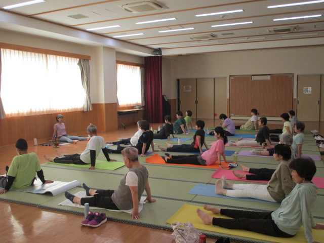 広々とした室内で、フローリングの上にヨガマットを敷いた多くの参加者が床に足を伸ばして座っており、正面にいる講師の女性の指示に従ってストレッチ運動をしている様子の写真