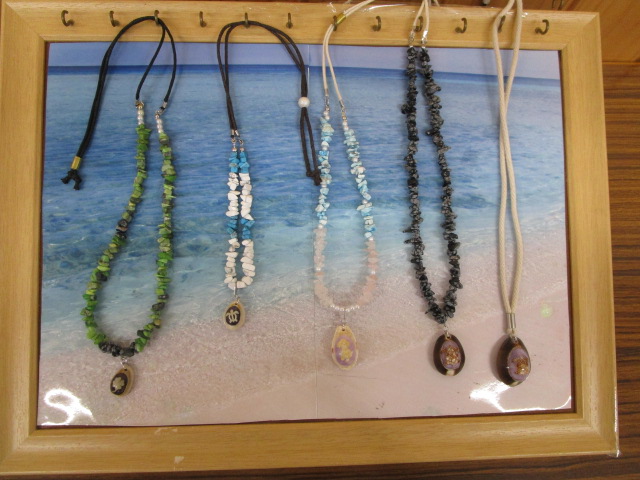 木製の額縁に、青い海辺の写真が背景として敷かれており、その上から小石状の天然石やビーズを使った手作りのネックレスが5本吊るされて飾られている写真