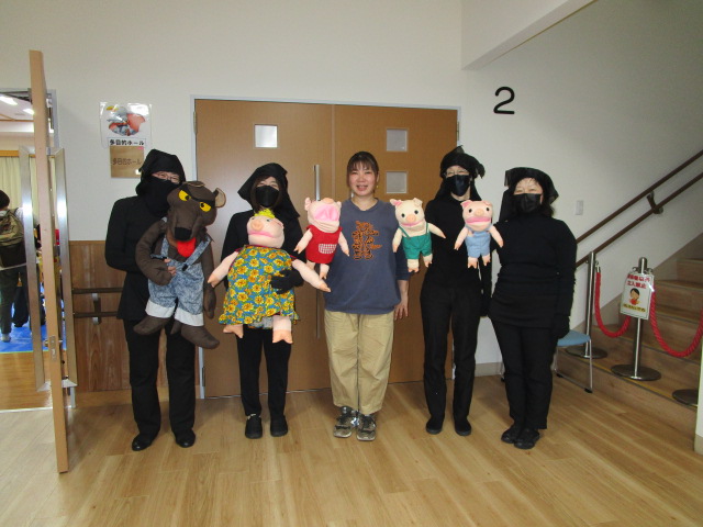 木目の床と白い壁の室内で、黒い服と頭巾で顔を隠した4人の人物と、私服の女性1人の計5人が並んで立っており、それぞれ「三びきのこぶた」をモチーフにした人形を持って笑顔でカメラを見ている写真