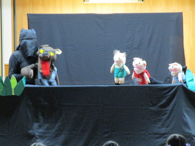 暗いステージの上で、黒い服を着て顔を隠した操作役の人物が、大きなオオカミの人形と3匹のこぶたの人形を使い、観客席にいる聴衆に向けて人形劇を披露している様子の写真