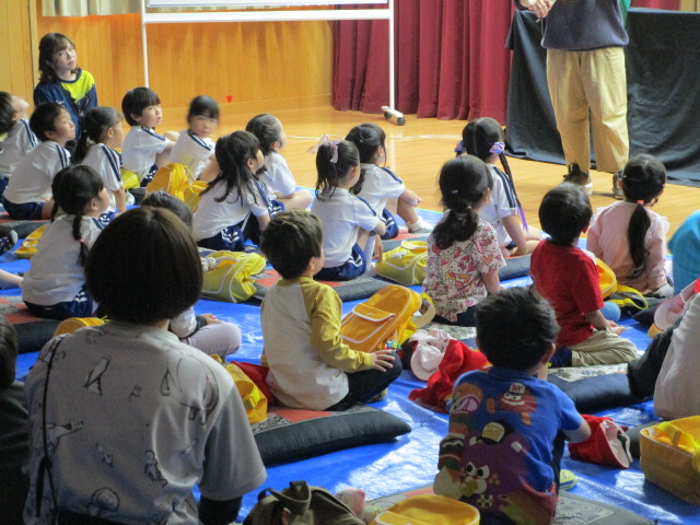 青いシートが敷かれた床に、多くの園児が座布団に座っており、正面の大人の話に熱心に耳を傾けている様子を後方から撮影した写真