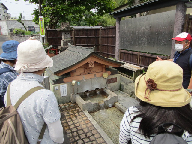 和風の小さな屋根がついた水汲み場で、数人の参加者が、赤い帽子をかぶった男性の説明を聞きながら、湧き水が流れ出ている様子を観察している写真