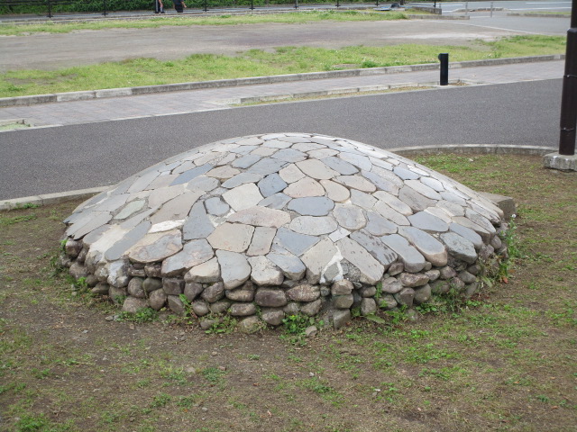 舗装された歩道に隣接する芝生の上に、丸いドーム状の石積み構造物が設置されており、表面と側面が自然な形や色合いの平たい石で丁寧に覆われている野外彫刻の写真