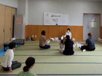 畳敷きの広い部屋で、数人の女性が床に座って向き合っており、中央の女性指導者が小さなタオルや布のようなものを使って、参加者に手遊びや交流の方法を指導している様子の写真