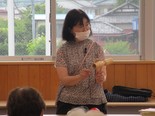 窓を背にした室内で、眼鏡とマスクを着用した講師の女性が、ウッドブロックを両手で持ち、座っている人たちに音を聞かせている様子の写真