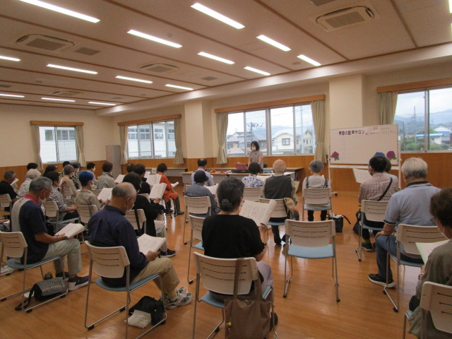 フローリングの広い室内で、白い椅子に座った多くの高齢者が楽譜を持ち、前方に立つ講師と思われる女性の指示を聞きながら歌の練習をしている歌声サロンの様子の写真