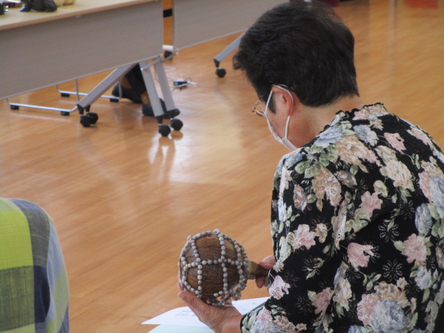 フローリングの室内で、花柄の服を着てマスクをした女性が、ビーズや貝殻のようなものが編み込まれた丸い形のヤシの実の楽器を持っている手元の写真