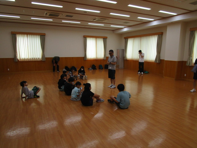 フローリングの広い室内で、中央に座って半円状に並んだ数人の子供たちに、半袖シャツの男性が立ち上がって何かを指導しており、他の大人は周囲で見守っている様子の写真
