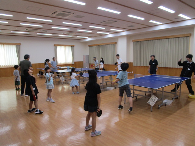 明るいフローリングの広い室内で複数の卓球台に、ラケットを持った子供たちが並び対面にいる大人に指導を受けている写真