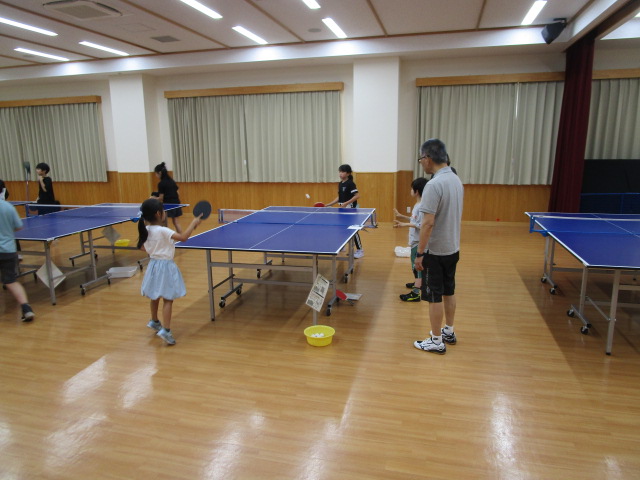 明るいフローリングの広い室内で複数の卓球台が設置されており、大人が見守る中、幼い女の子が試合形式の練習をしている様子の写真