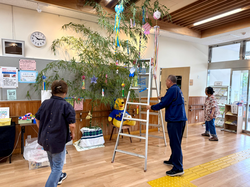 明るい室内の広間に、大きな笹飾りが立てられており、脚立に登った男性が笹に飾りを吊るしている様子を、他の参加者と思われる2人の女性が手伝いながら見ている、七夕の飾り付け準備の様子の写真