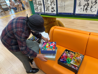 オレンジ色のソファに置かれた箱の中のカラフルな折り紙の飾りを、チェック柄のシャツと黒い帽子をかぶった年配の女性が身をかがめて触って確認している写真