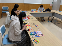 明るい会議室のような部屋で、長机をコの字型に囲んで3人の女性が椅子に座り、紙や布、ハサミ、接着剤などを使って様々な形に切り貼りしている写真
