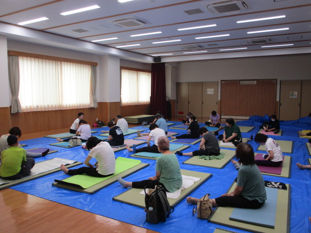 フローリングの広い室内で、青いビニールシートの上にヨガマットを敷いた多くの参加者が座って開脚のストレッチをしている写真